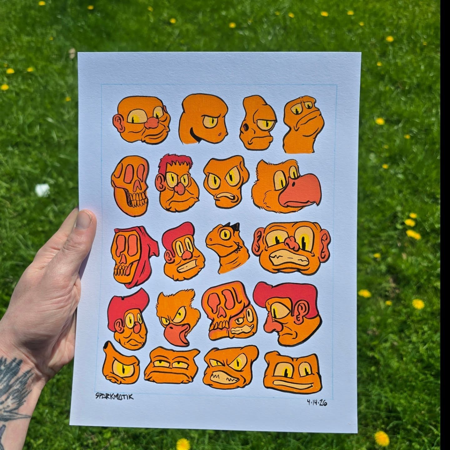Twenty small orange cartoon faces — skulls, goblins, a bird, a lizard — painted in rows on white paper, signed sparKmatiK.