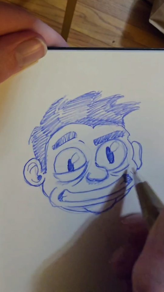 Loose blue ballpoint pen sketch of a grinning guy with scribbled hair and big mismatched eyes in a sketchbook.
