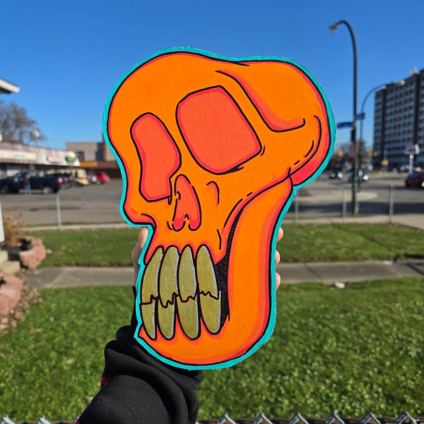 Hand-cut wood skull painted hot orange with gold teeth and a teal outline, held up against a blue Buffalo sky.