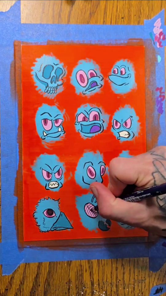 Grid of fuzzy blue monster faces with pink eyes — including a skull and a bird — painted on a bright orange-red panel taped to the table.