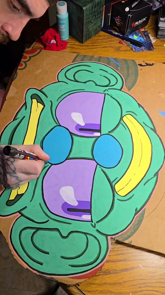 Huge green monster head with purple eyes and yellow bananas for ears, painted in acrylic marker on a large sheet of found cardboard.